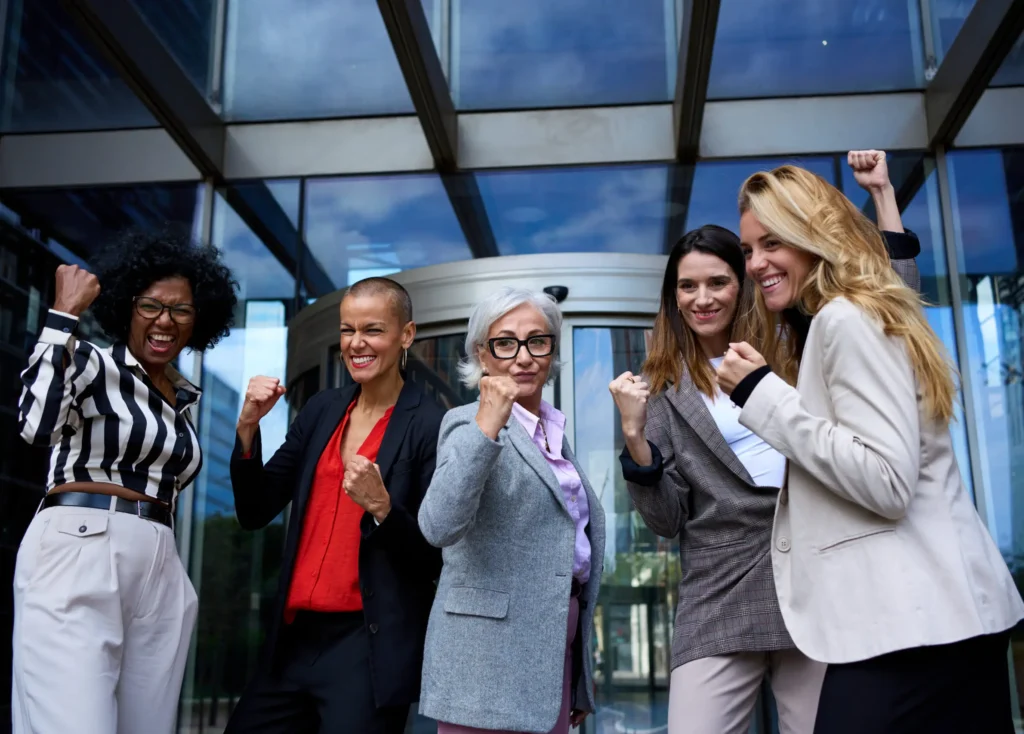 Five women stand together outside a glass building entrance, smiling and flexing their arms in a gesture of strength and celebration. They are dressed in business attire, exuding confidence and camaraderie.