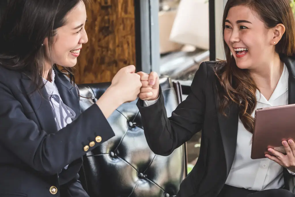 Two women in business attire are smiling and giving each other a fist bump while sitting on a leather couch. One woman holds a tablet. They appear to be in a positive and friendly discussion about strategic growth in their field.