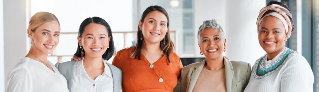 Five women, standing closely together and smiling at the camera, in a well-lit indoor setting. Dressed in colorful and professional attire, they showcase diversity and camaraderie. The background features modern office furniture, highlighting a successful women-led business environment.