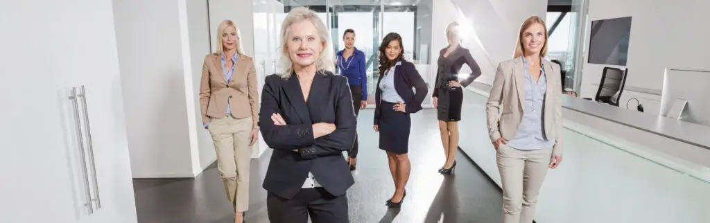 A group of six professionally dressed women stands confidently in a modern office space. The woman in the front crosses her arms, while the others pose dynamically behind her, creating an empowering scene that epitomizes Women-Led Startup Success.