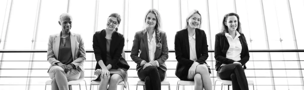 Black and white depiction of five women sitting on chairs in a row, all wearing business attire and smiling. They are seated in front of a large window that provides a bright, soft-lit background with metal bars visible through the glass. A testament to Women-Owned Business Success.