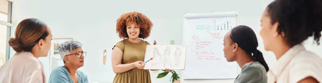 A woman stands at the head of a table, holding a drawing. She is presenting to four people seated around the table, discussing strategies for revenue acceleration. A flipchart with diagrams and notes is visible in the background. The setting appears to be a professional meeting or brainstorming session.