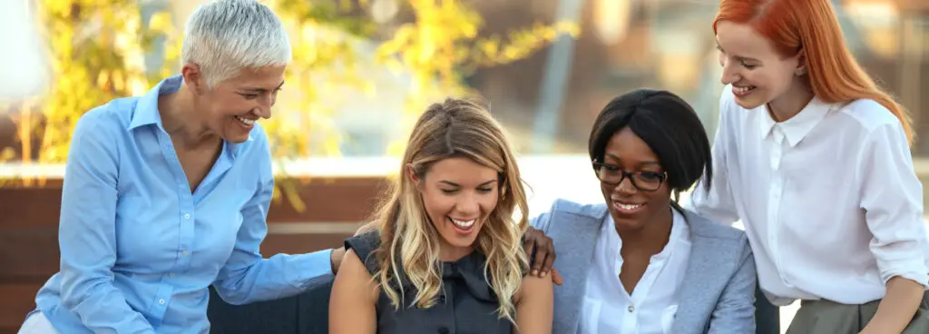 Four women are gathered around a laptop, smiling and engaged in a discussion outdoors, embodying the spirit of Women-Owned Business Success. One woman is seated with the laptop, while the others stand or kneel around her. The background features blurred greenery and a building.