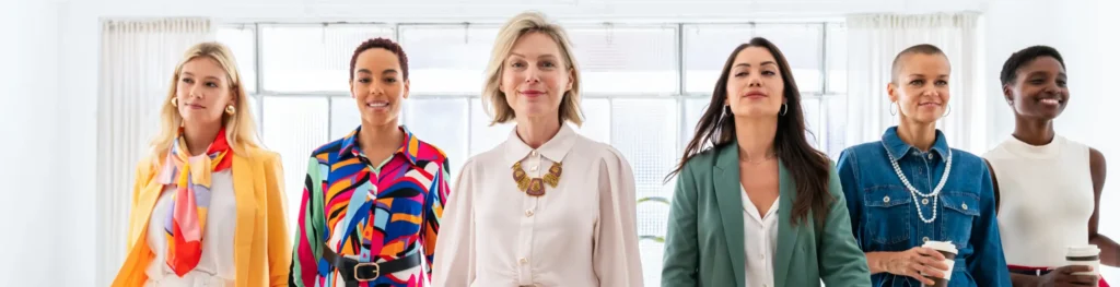 A diverse group of six women confidently walks forward in a well-lit room. They are dressed in a variety of colorful and stylish outfits, with some holding documents and others holding coffee cups. The atmosphere of this women-led startup success appears professional and empowering.