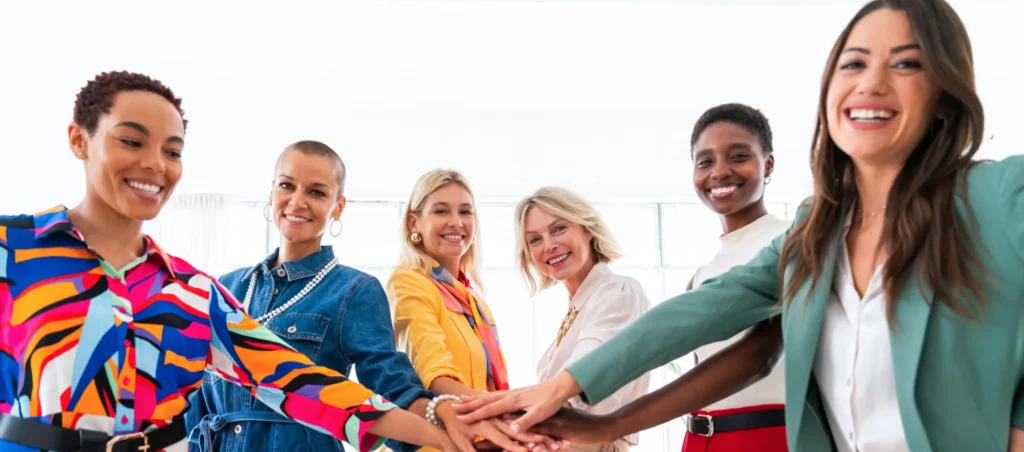 A group of six diverse women stand together in a line, smiling broadly, and stacking their hands in the center. They are dressed in bright, colorful clothing, embodying Female Business Leaders and suggesting a cheerful and collaborative atmosphere focused on Empowering Women in Business.