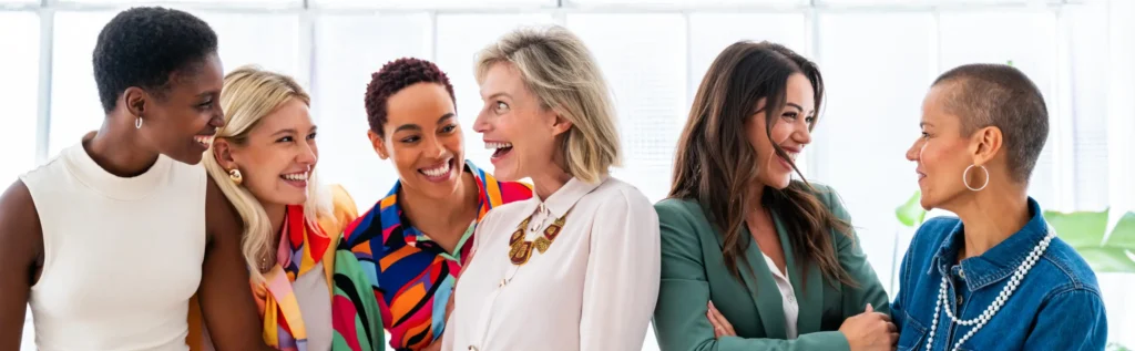 A group of six women stand together in a well-lit room, smiling and laughing. They are dressed in vibrant, stylish clothing and appear to be enjoying each other's company. The background features large windows, allowing natural light to flood in, creating a perfect setting for thriving women-led companies.