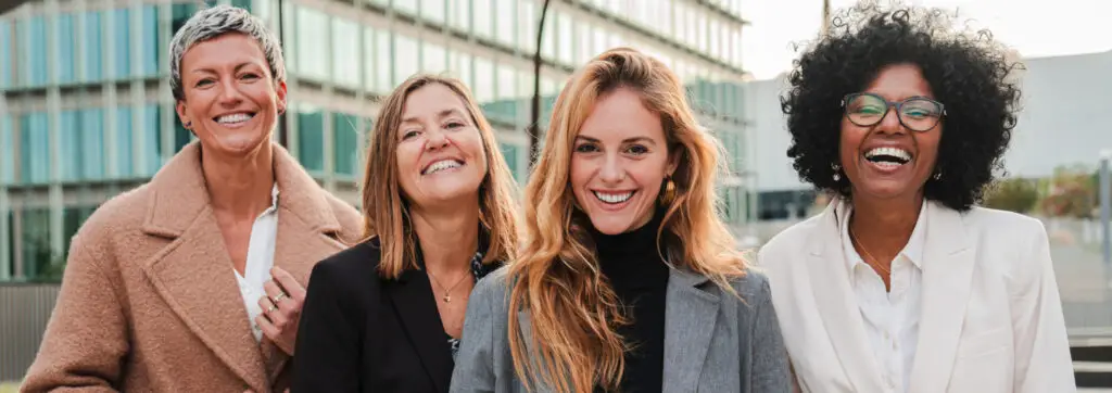 Four business women dressed in professional attire stand together outside a modern glass building, smiling warmly at the camera. Three women are in suits, and the fourth is in a long coat. Their expressions suggest camaraderie and confidence, embodying women leadership and empowerment in a professional setting.