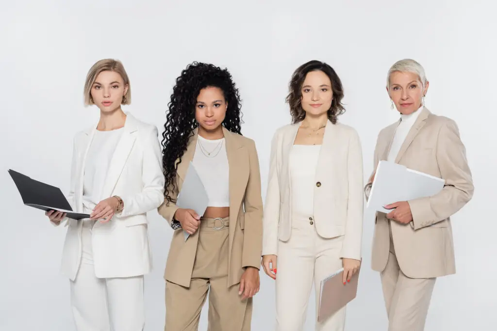 Four women of diverse appearances stand confidently in a row, all dressed in stylish, professional business attire. Each holds electronic devices or folders, indicating a work setting. The plain white background underscores their professionalism and unity, showcasing empowering women in business.