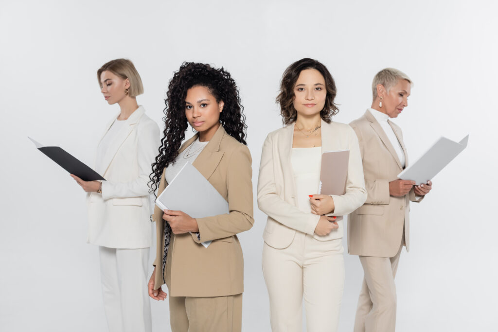 Four professionally dressed individuals stand against a plain background. Three hold folders while one holds a tablet. Two face forward, and the other two stand back-to-back, looking at their documents. They appear focused and engaged in their tasks, embodying empowering women in business with determination and expertise.