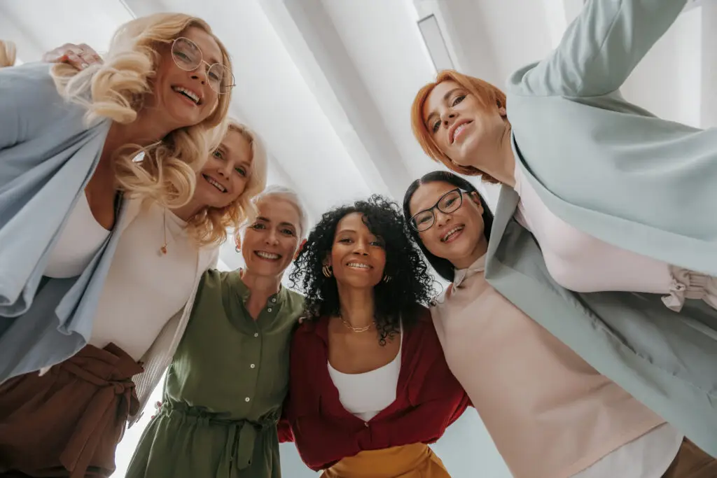 A group of six diverse women stand together in a circle, smiling and looking down at the camera. Dressed in colorful, stylish clothes, they appear happy and connected in the bright, well-lit room—an inspiring portrait of female business leaders celebrating their success.