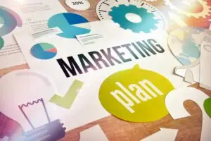 A colorful and cluttered workspace filled with marketing-related elements. The word "MARKETING" in bold black letters is prominent, with the word "plan" in green over a speech bubble. Various charts, gears, and icons surround the main text.