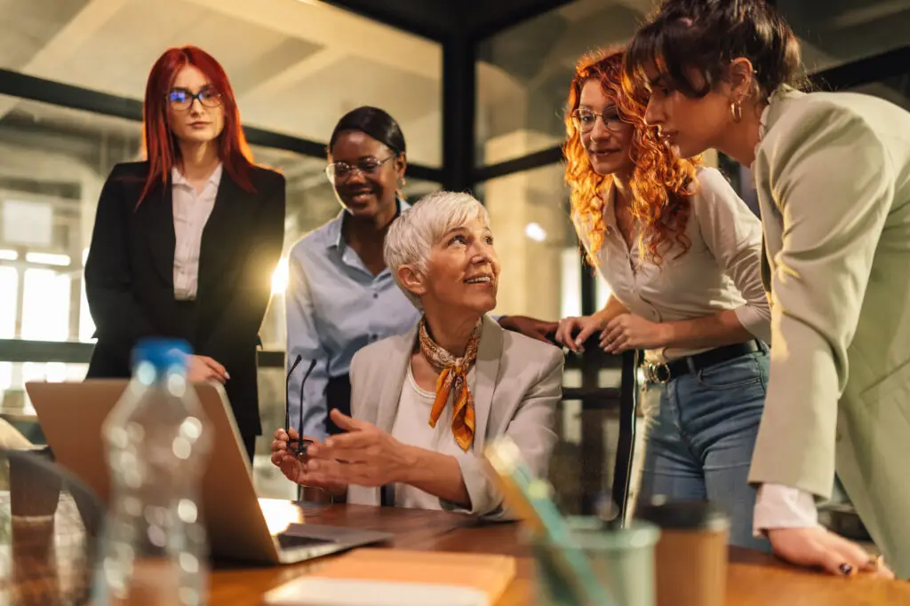 An older woman with short white hair, dressed in a blazer, is sitting at a table with an open laptop and discussing something with four women standing around her. In a modern office with glass walls, these female business leaders appear engaged and collaborative.