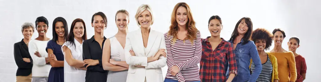 A group of thirteen diverse women stand in a V-formation, smiling confidently. They are dressed in various casual to business-casual outfits and are situated in a bright room with a white brick wall. Exemplifying empowering women in business, the women in front have their arms crossed or hands on hips.