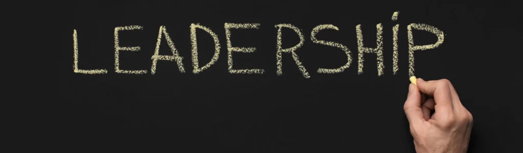 A person's hand, embodying the essence of Empowering Women in Business, writes the word "LEADERSHIP" in large, capital letters with yellow chalk on a blackboard. The background is a solid dark color, and the word is centered and underlined by the person's left hand holding the chalk.