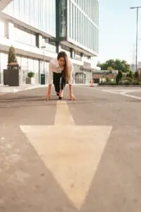 A woman in business attire crouches in a runner's starting position on an arrow painted on an empty urban street. She appears focused and ready to sprint, with modern buildings and greenery in the background on a bright, sunny day.