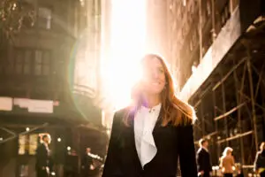 A woman with long hair, wearing a white blouse and dark blazer, smiles in a sunlit urban setting. Sunlight shines brightly behind her, casting a halo effect. People walk around in the background, surrounded by tall buildings and scaffolding.