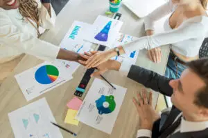 Three colleagues seated around a table with charts and graphs, placing their hands together in the center as a gesture of teamwork. The table is covered with various business documents and a few stationery items.