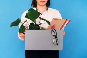 Person holding a box containing a potted plant, a pair of glasses, stationery, and files, standing against a blue background. The person is partially visible from the neck down, wearing a white shirt.