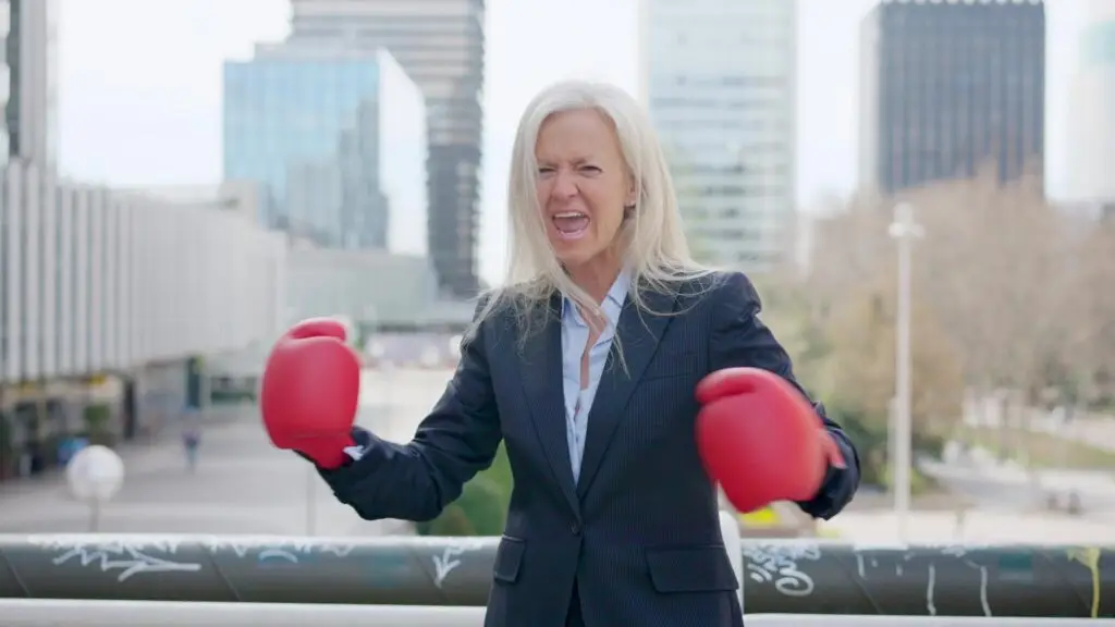 A woman in a navy business suit wears red boxing gloves and stands outdoors in an urban setting with tall buildings in the background. As a certified resilience coach, she has a determined expression with her mouth open and fists raised.