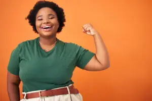 A person with short curly hair, wearing a green t-shirt and beige pants, smiles confidently while flexing their arm in front of an orange background. They are accessorized with necklaces and a brown belt.