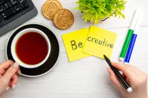 A person writes "Be creative" on yellow sticky notes beside a cup of tea, cookies, a small green plant, and a keyboard. Markers in green, blue, and black are placed nearby on a white wooden desk.