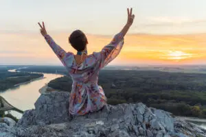 A person wearing a floral dress sits on a rocky cliff with their back to the camera, arms raised in a peace sign. They overlook a scenic sunset with a river winding through a forested landscape below.